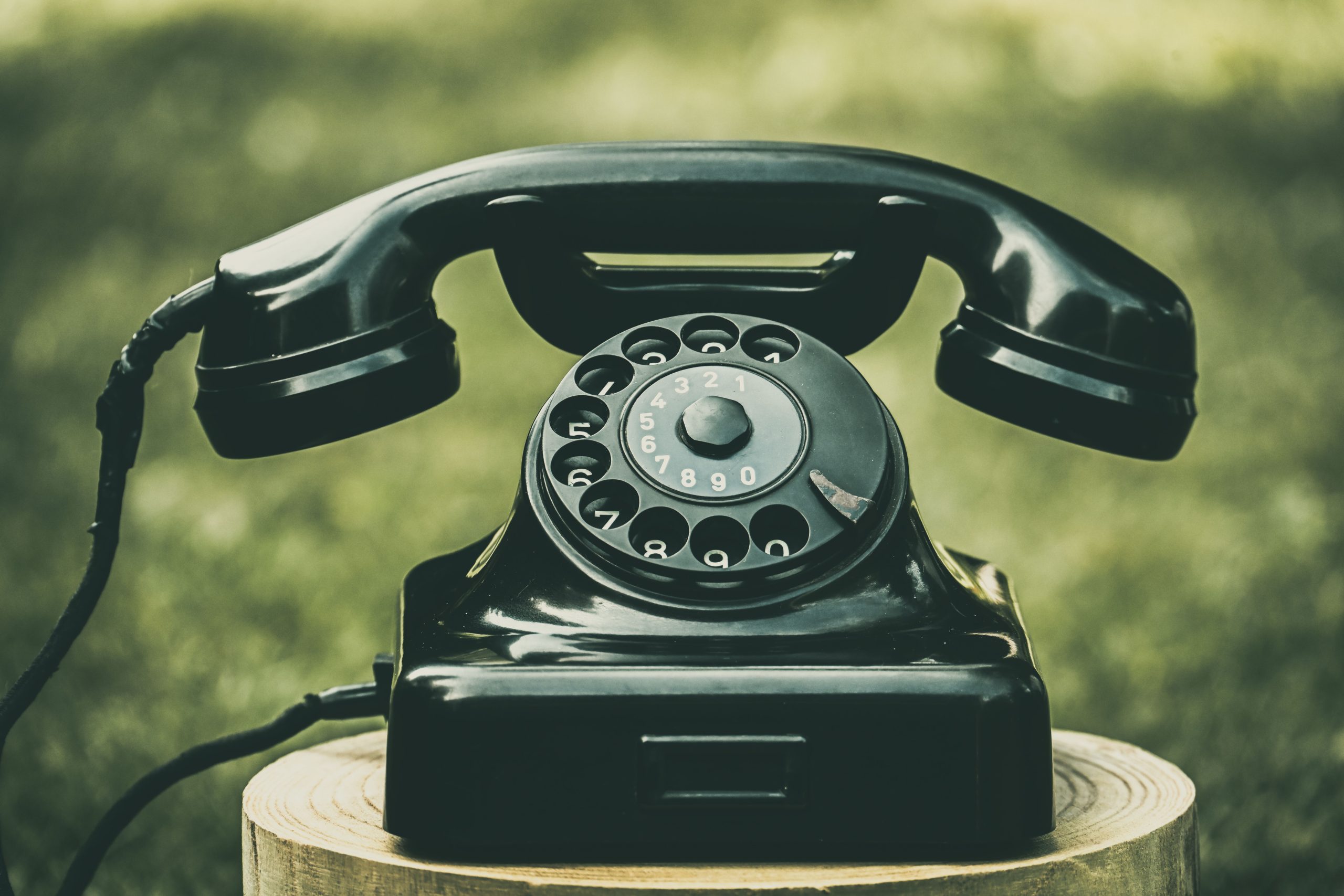 A classic black rotary dial telephone placed outdoors on a wooden stump, evoking nostalgia.